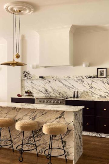 Modern kitchen featuring a marble countertop, gold pendant light, and plush bar stools, with dark cabinetry and stainless steel appliances.