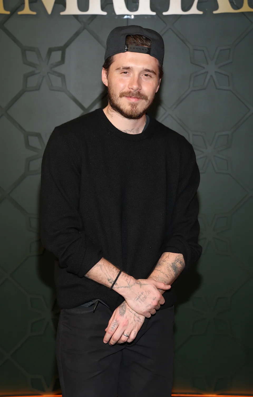 Brooklyn Beckham wearing a casual sweater and cap stands with hands crossed, showcasing tattoos on arms