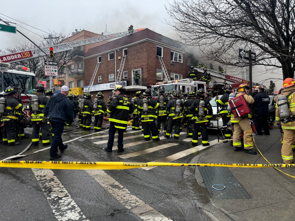 Firefighters battling a fire at a three-story multiple dwelling, with smoke billowing from the second floor windows and roof.