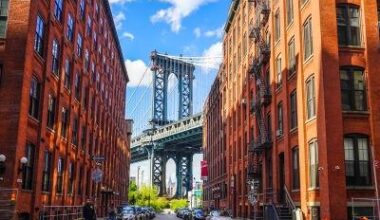 This iconic urban landscape captures the historic DUMBO neighborhood in Brooklyn, framed by the classic red-brick architecture of former industrial warehouses. The view centers on the massive steel towers of the Manhattan Bridge, perfectly aligned between
