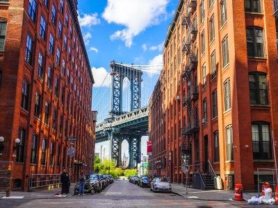 This iconic urban landscape captures the historic DUMBO neighborhood in Brooklyn, framed by the classic red-brick architecture of former industrial warehouses. The view centers on the massive steel towers of the Manhattan Bridge, perfectly aligned between