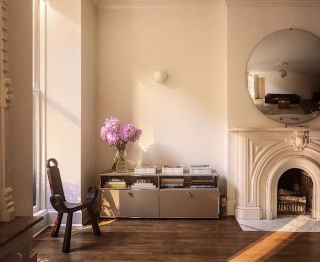 A corner of a living room featuring a black chair, a vase of pink flowers, a mirrored wall, and a marble fireplace.