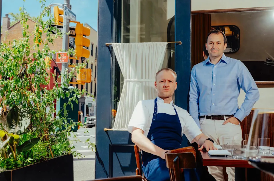 One man in an apron sitting at the table with another man in a blue shirt standing next to him.