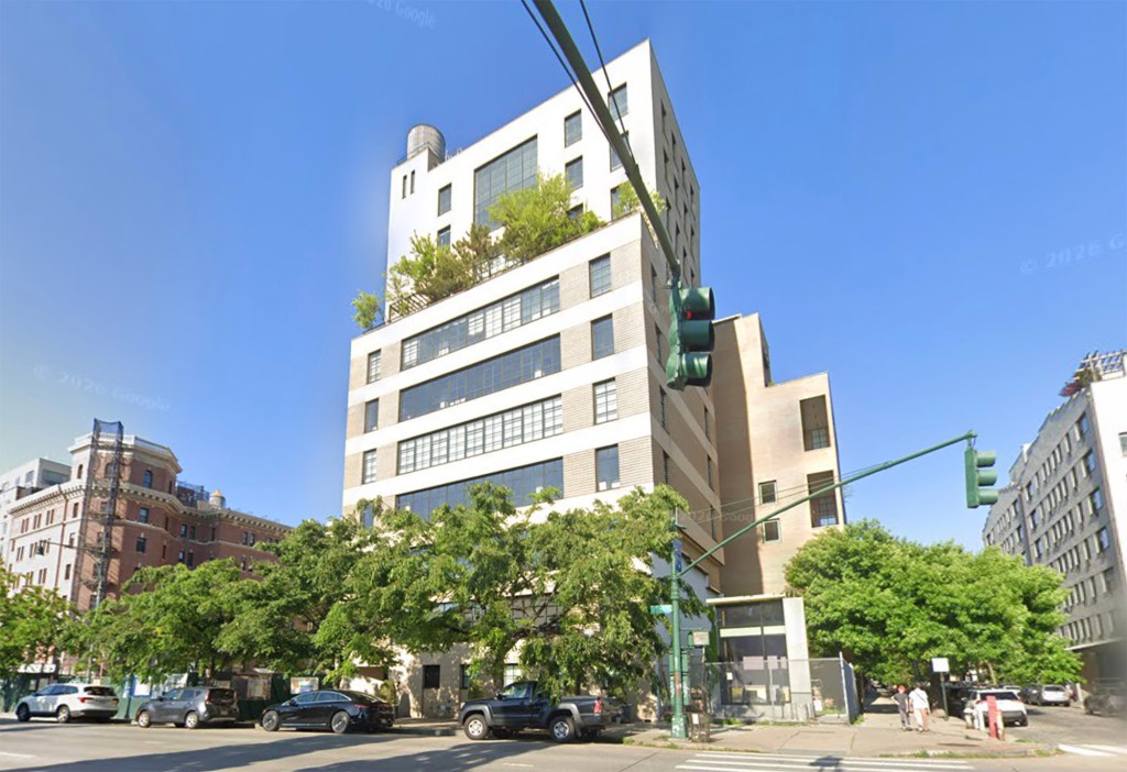 A tall, white apartment building with a water tower on top, balconies with trees, and large windows, on a street with cars and other buildings.