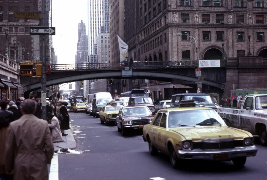 Busy city street scene with several yellow taxis, pedestrians on sidewalks, and tall buildings. An overpass bridge crosses above the traffic. The setting appears to be in New York City.
