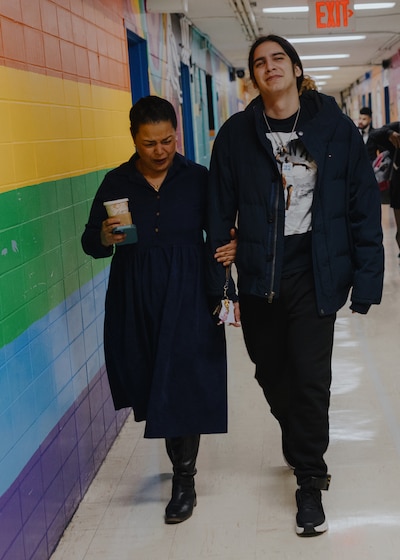 A photograph of an adult woman holding onto the arm of an adult high school student while walking down a school hallway.