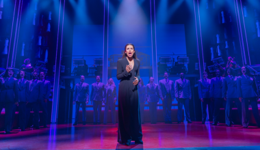 A woman stands and sings at center stage in dramatic blue and purple lighting, with a chorus of people in suits standing behind her on a theater set.