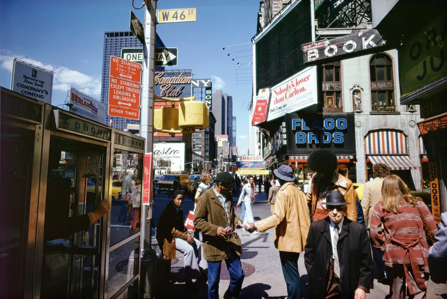 A busy city street scene with people walking, colorful storefronts, neon signs, a phone booth, and street signs, including W 46th St. The scene appears lively, capturing a moment from decades past.