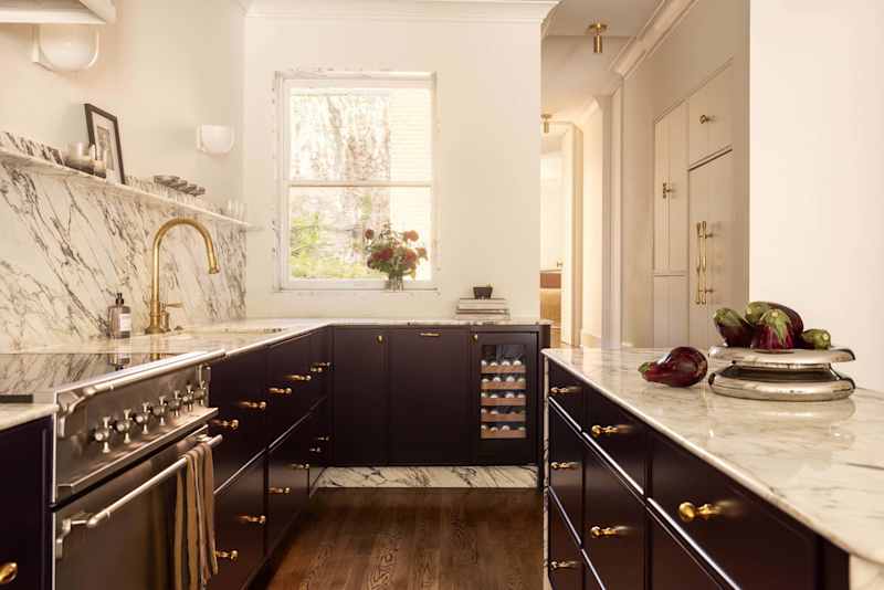 Modern kitchen with dark cabinetry, marble countertops, a gold faucet, and a window with a floral arrangement.