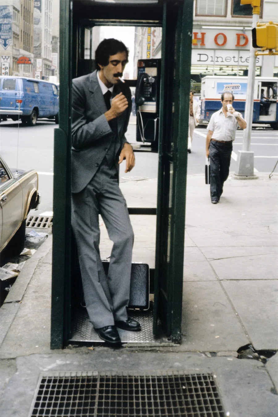 A man in a gray suit stands in a phone booth on a city sidewalk, holding the phone to his ear. A briefcase is at his feet, and people and cars are visible on the busy street in the background.