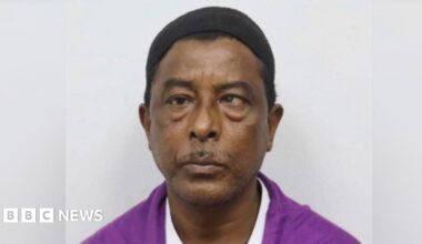 Headshot of a man looking straight ahead with a small black cap on his head, wearing a magenta shirt