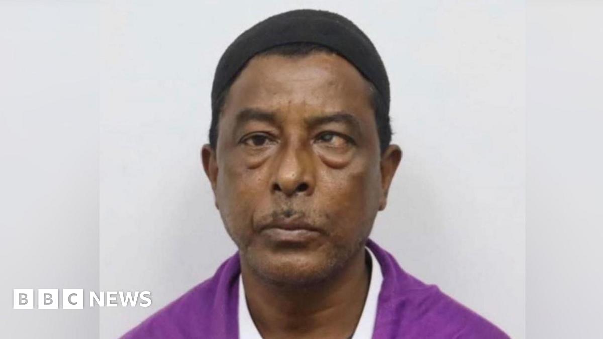 Headshot of a man looking straight ahead with a small black cap on his head, wearing a magenta shirt