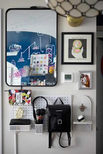 A wall display featuring a mirror with colorful doodles, framed art, a thermostat, and organized accessories like a backpack, headphones, and pens.