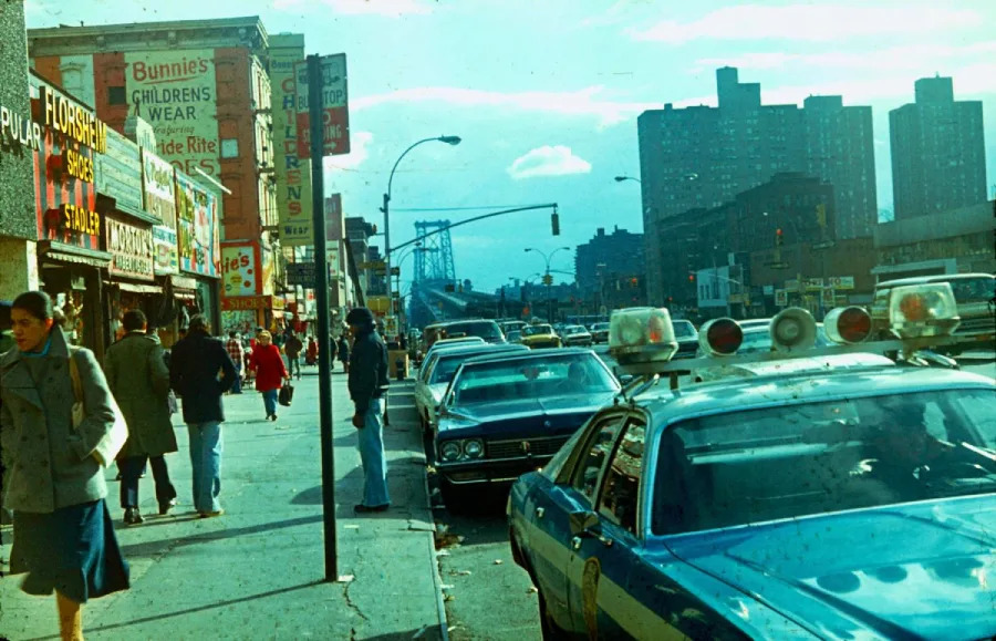 A busy city street scene from the 1970s shows pedestrians walking past parked cars and a police car. Storefronts with colorful signs line the sidewalk, and tall buildings and a bridge are visible in the background.