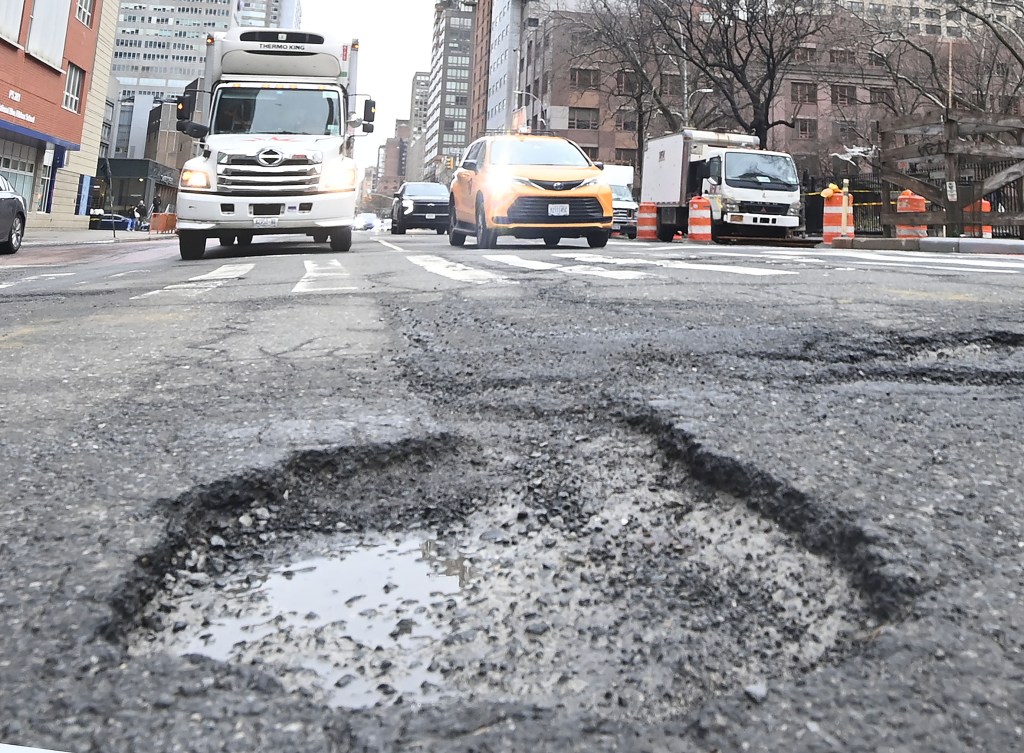 A large pothole with standing water in the foreground, with traffic and buildings in the background.