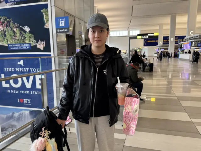 A woman in a grey baseball hat and black jacket holds luggage and a pink drink while standing in an airport concourse
