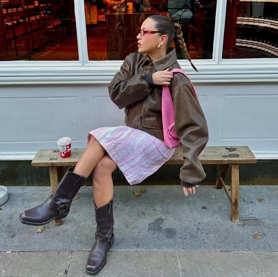  A woman wearing a brown leather jacket, pink shoulder bag, pink striped skirt, and brown boots. 