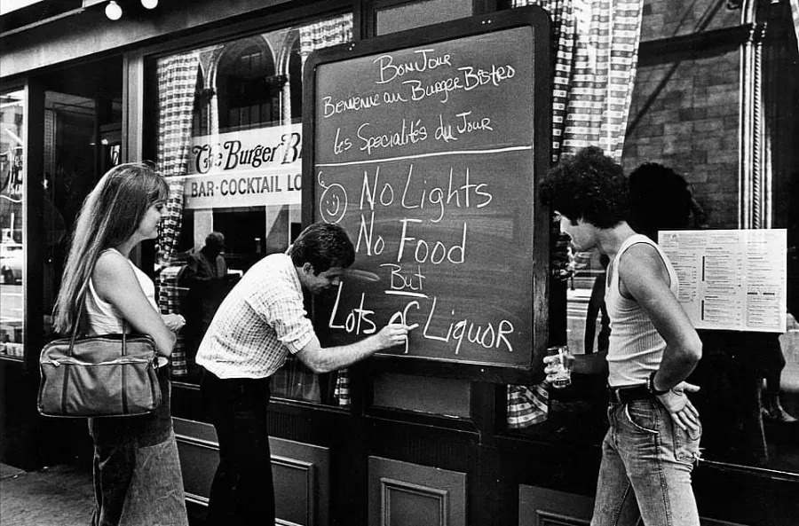 Three people stand outside a restaurant as one writes on a large chalkboard sign that humorously reads: “No lights, No food, But lots of liquor.” Two people watch and smile. The scene is set on a city street.