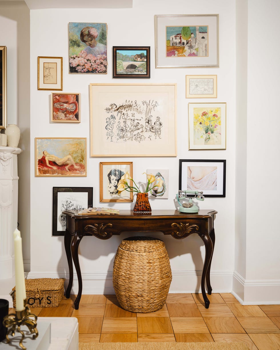 Wall with various framed artworks above a wooden console table and woven stool.
