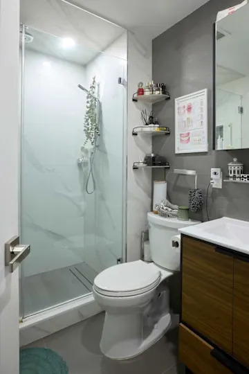 Modern bathroom featuring a glass shower, toilet, and wooden vanity with storage. Shelves hold toiletries and decor.