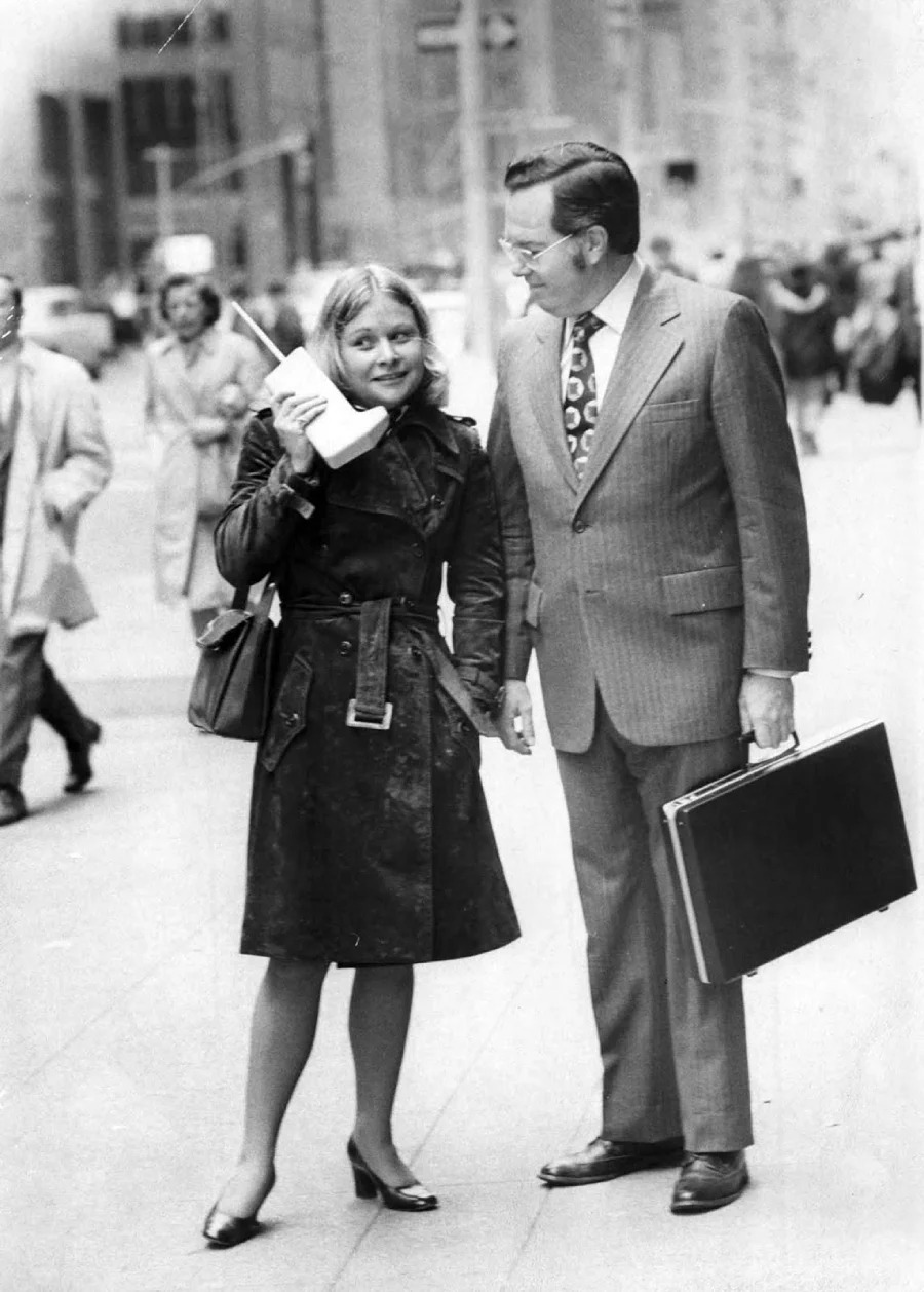 A woman in a trench coat talks on an early, large mobile phone while standing on a city sidewalk next to a man in a suit holding a briefcase. Several pedestrians walk by in the background.