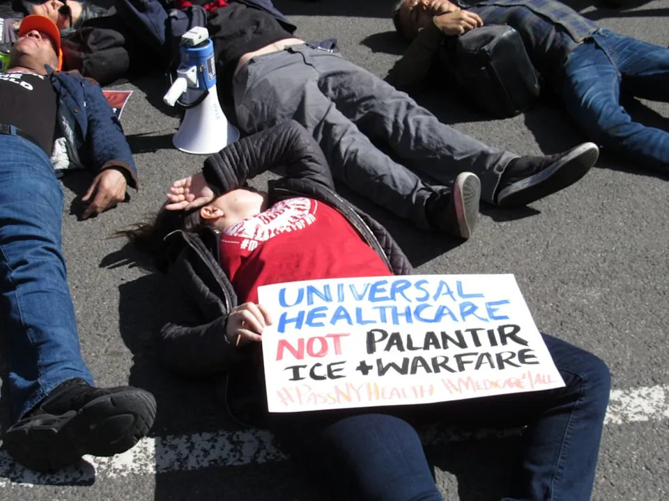 People lie in a city street. One at the center of the photo lies with a hand covering their face, holding a sign in their other hand that reads: "Universal health care, not Palantir, ICE & warfare."