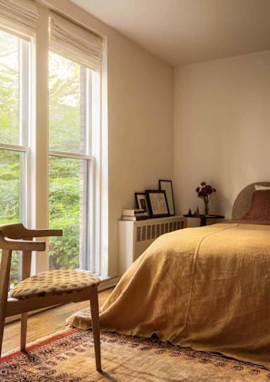 Cozy bedroom with a mustard-colored bedspread, wooden chair, and large windows overlooking greenery.