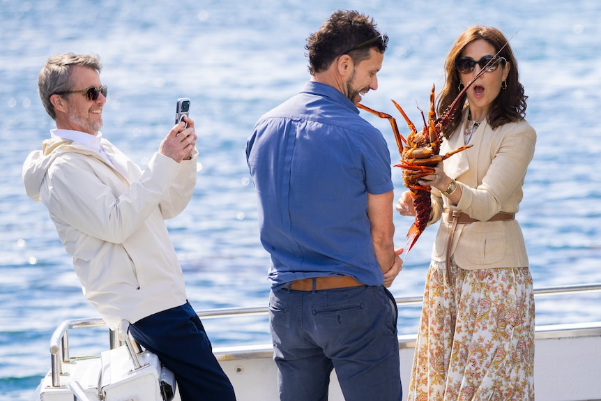 King Frederik takes a photo of Queen Mary holding a lobster while they are on a boat.