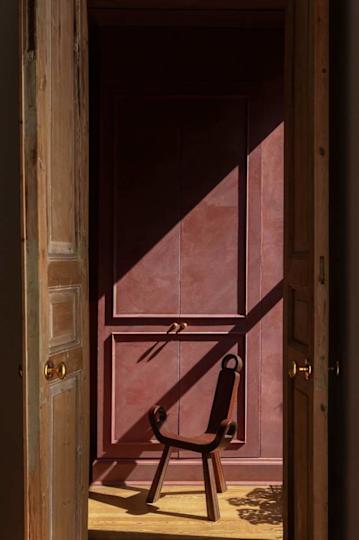 A wooden chair with curved arms sits in a softly lit entryway, framed by two doors and a textured red wall.