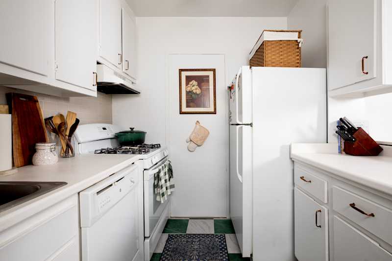 Narrow kitchen with white cabinets, green pot on stove, wooden utensils, and a patterned rug on the floor.