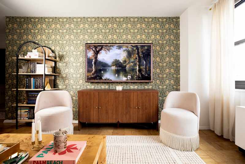Cozy living room featuring two cream-colored chairs, a wooden sideboard, and a landscape painting on patterned wallpaper.