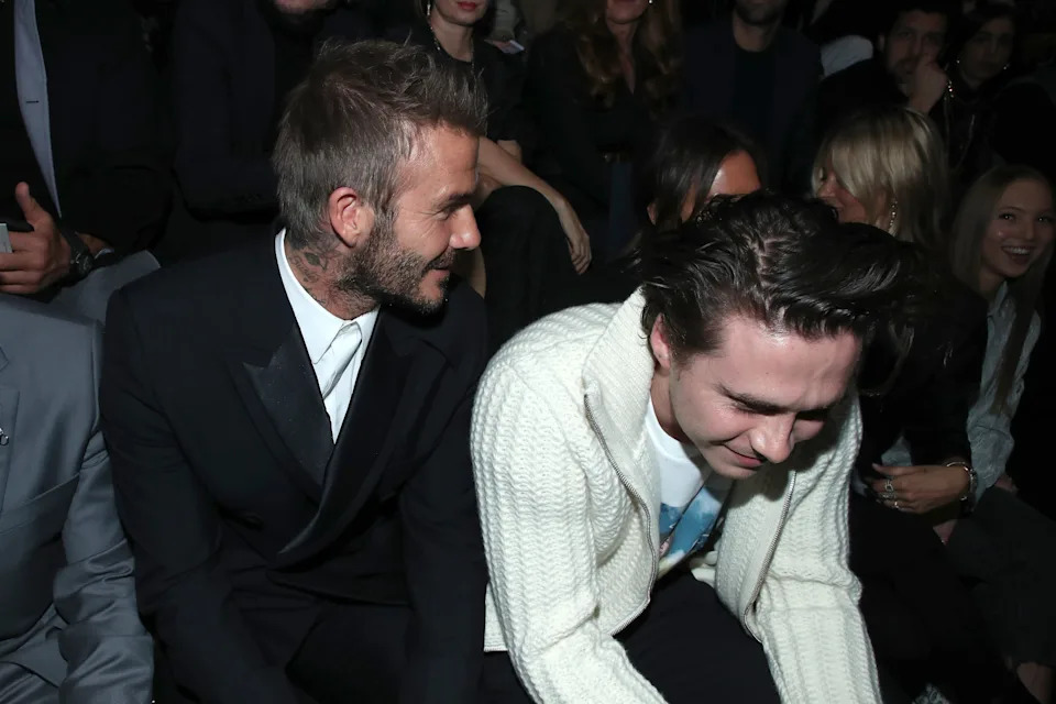 David and Brooklyn Beckham seated at an event, both smiling and engaging with each other
