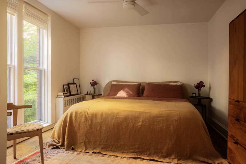 Cozy bedroom with a mustard-colored blanket, two pink pillows, a wooden chair, and a small side table with flowers.