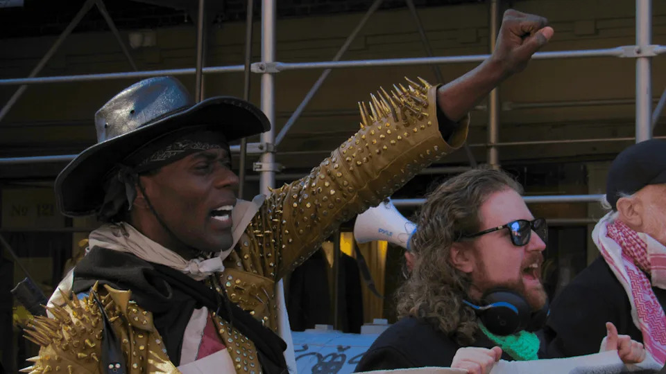 A person wearing a gold, spiky jacket and black cowboy hat raises a fist in the air and appears to be calling out something.