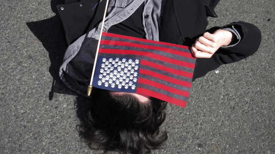 A person lies in the street and holds a black and red American flag over their face.