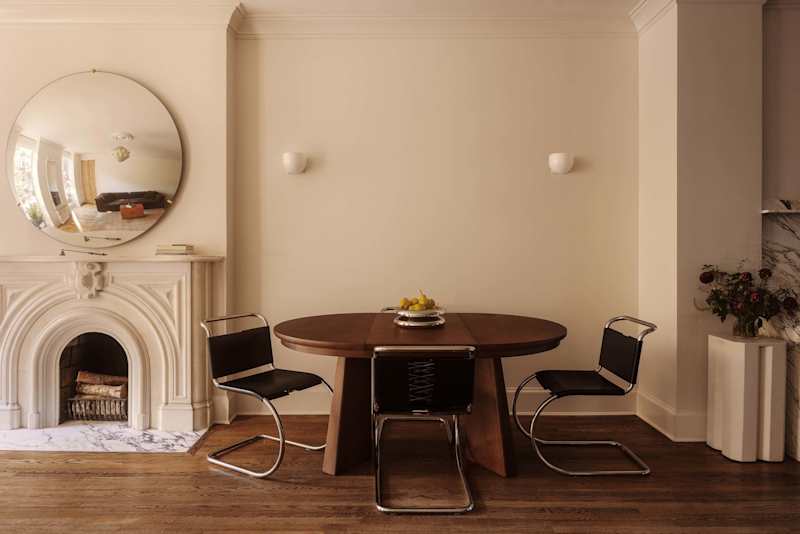 Round wooden dining table with a fruit bowl, surrounded by black chairs, near a marble fireplace and a large mirror.