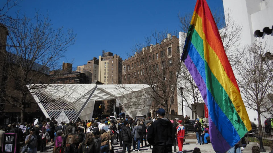 A person holds a large Pride flag outside of a cluster of people holding picket signs in front of the white New York City AIDS Memorial.