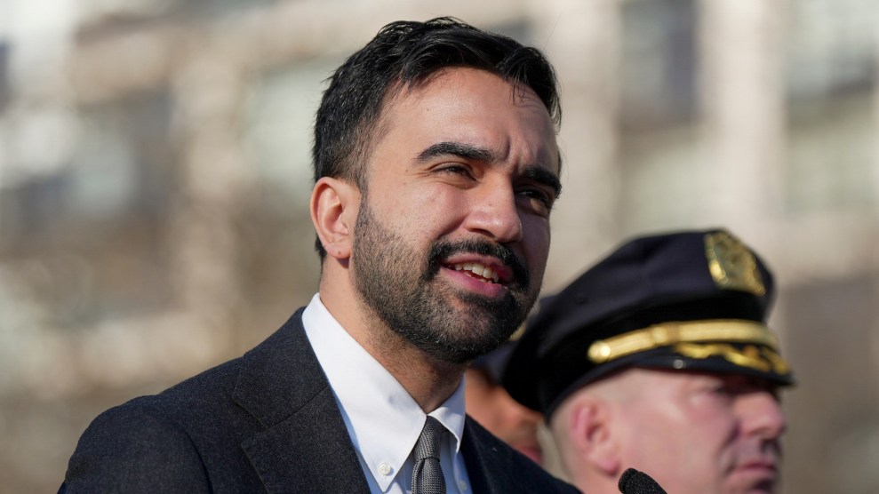 Zohran Mamdani, dressed in a black suit, stands outside and speaks into a microphone. A police officer, wearing a police cap, is visible in the background standing next to Mamdani.