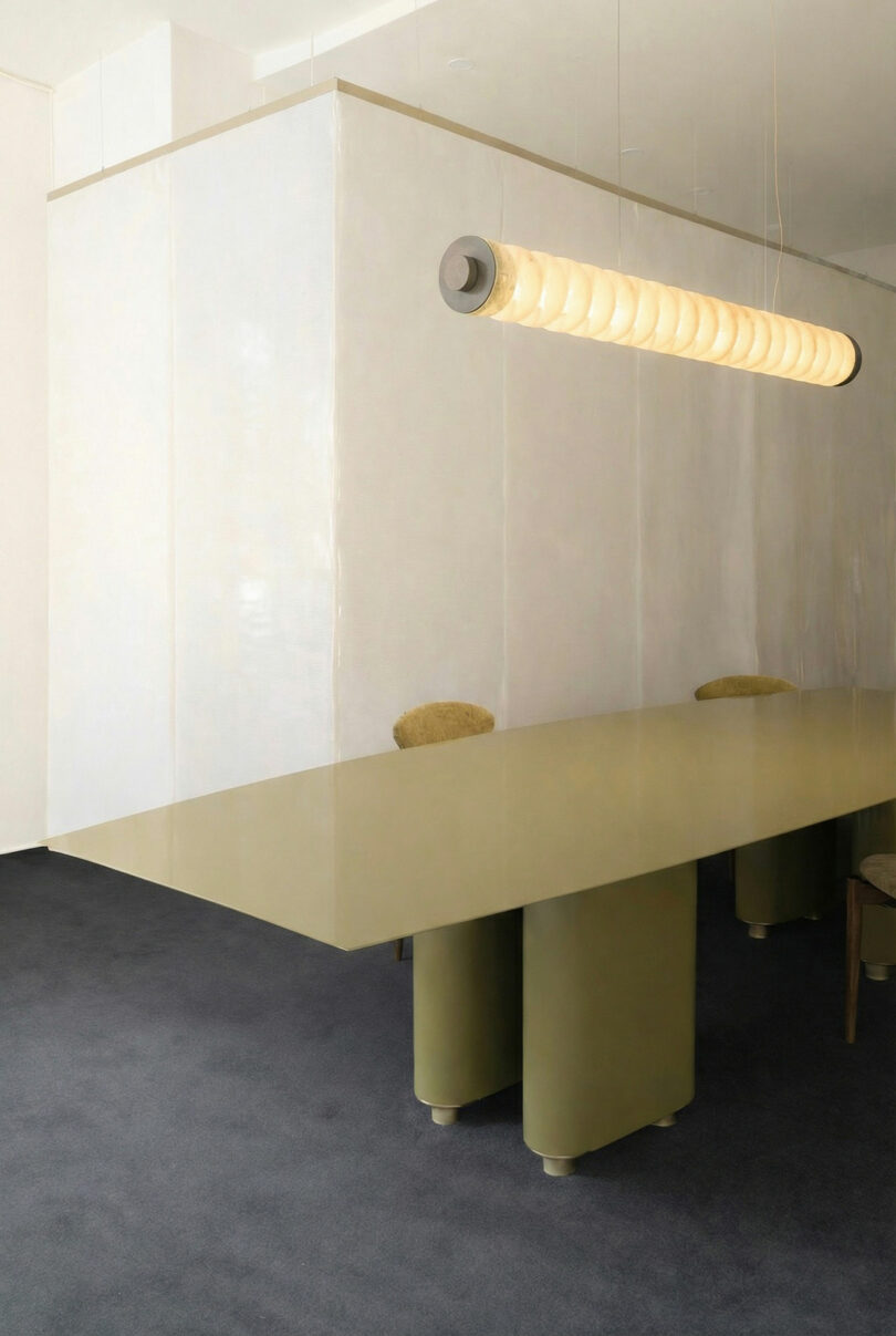 Minimalist meeting room featuring an ATRA large olive-green table, beige chairs, and a long cylindrical ceiling light fixture, set against light-colored walls and a dark carpeted floor.