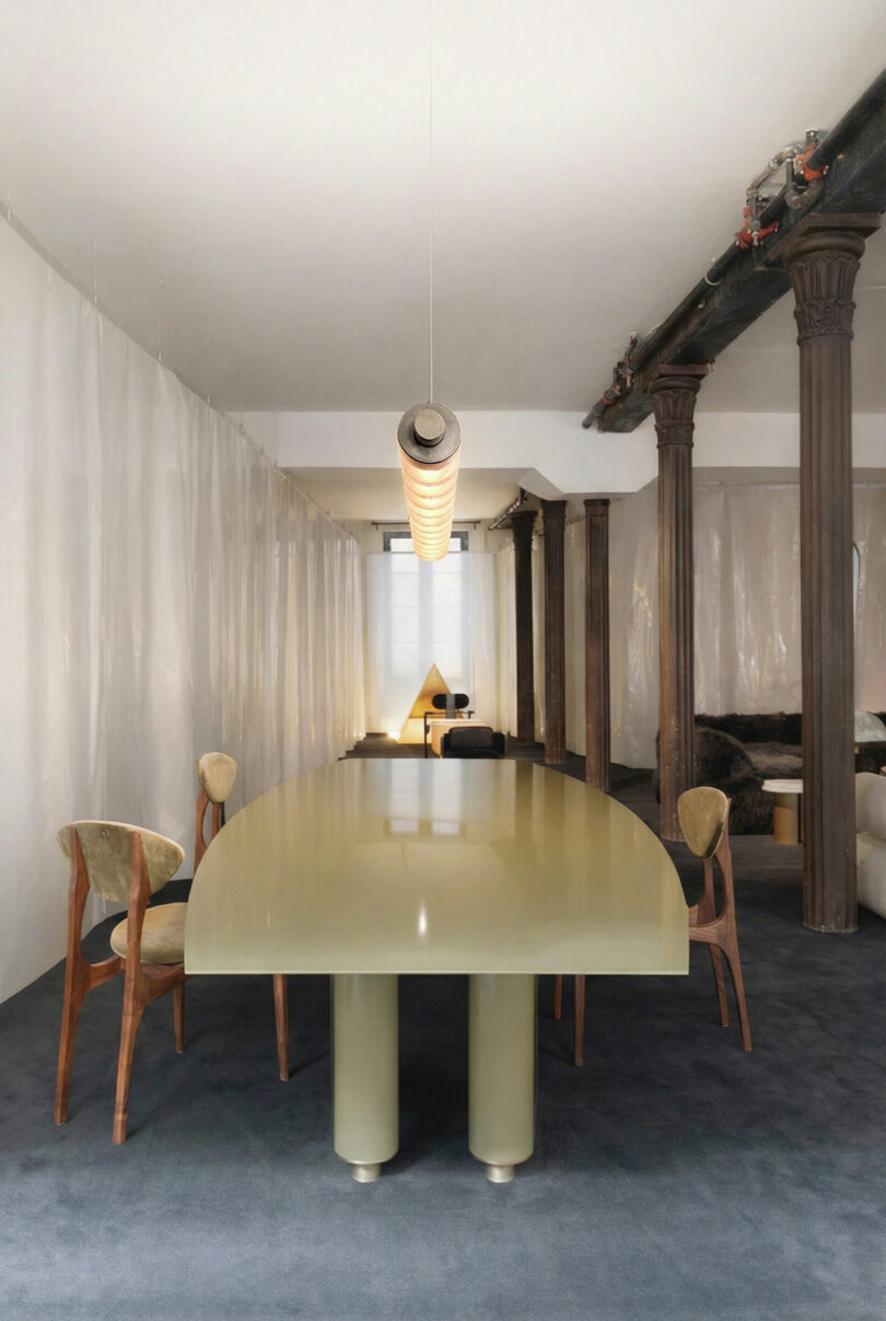 A modern dining room featuring a glossy, curved rectangular ATRA table, four wooden chairs, exposed pipes, and tall columns on a dark carpeted floor. Translucent curtains line the walls.