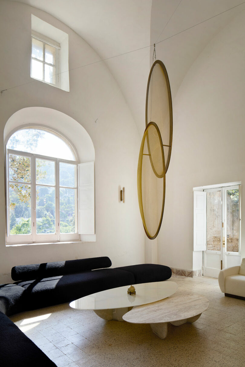 A modern living room with ATRA curved black sofas, a white round coffee table, large arched window, and two overlapping circular sculptures suspended from the ceiling.
