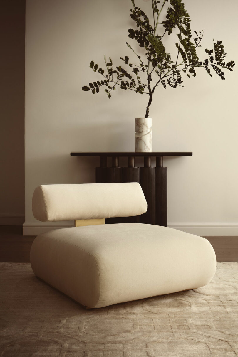 A modern beige lounge chair by ATRA sits on a textured rug in front of a dark console table with a marble vase holding green branches.