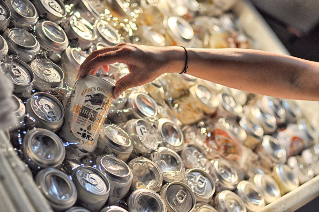 A person's hand reaches into a cooler filled with ice and many aluminum cans, pulling out a can of Kirin Beer.