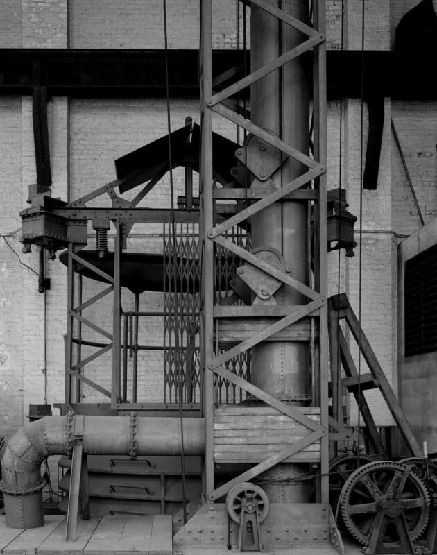 Black and white photo of industrial machinery, featuring metal beams, gears, pipes, and a lattice tower structure, set against a brick wall inside a factory or workshop.