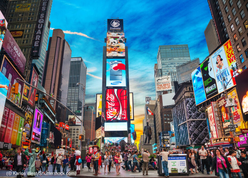 Tourists visiting Times Square walk below brightly lit billboards (© Karlos West/Shutterstock.com)