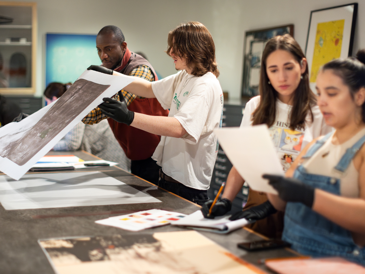 Students wearing protective gloves examine large prints spread across a table in an art study room.