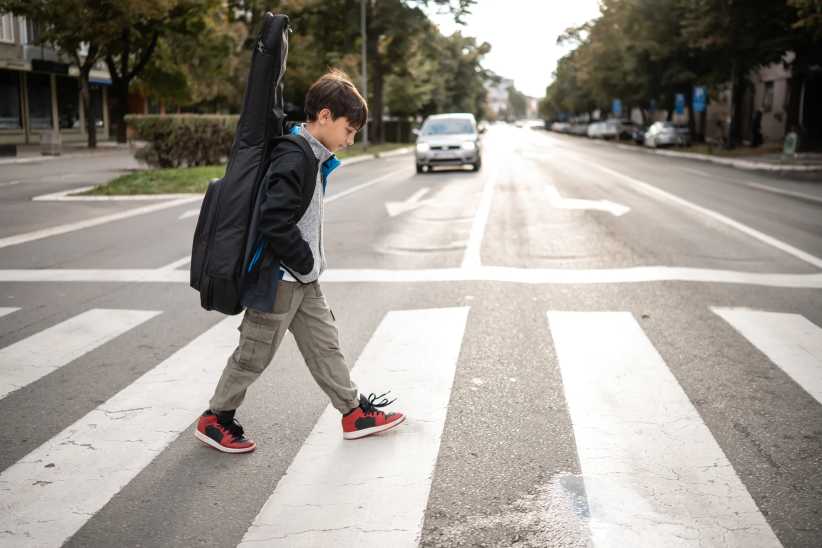 NYC Is Dropping Speed Limits to 15 MPH Near Every School Young boy walks across a city crosswalk carrying a guitar bag, heading to music lesson or school amid urban buildings and traffic