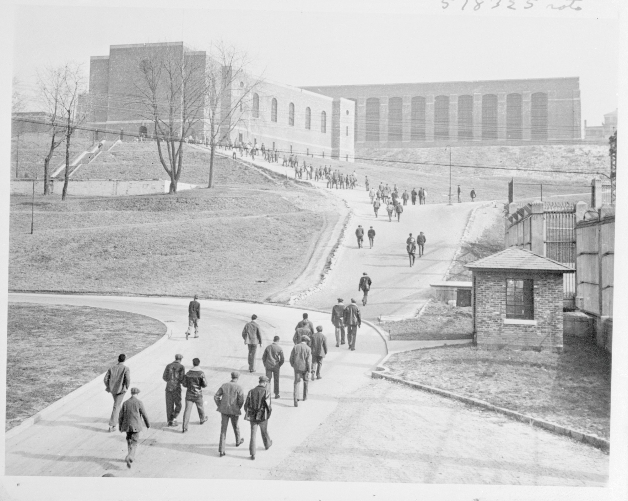 Groups of people walk along pathways through a large institutional compound with brick buildings, earthen embankments, and fenced areas on a barren landscape.