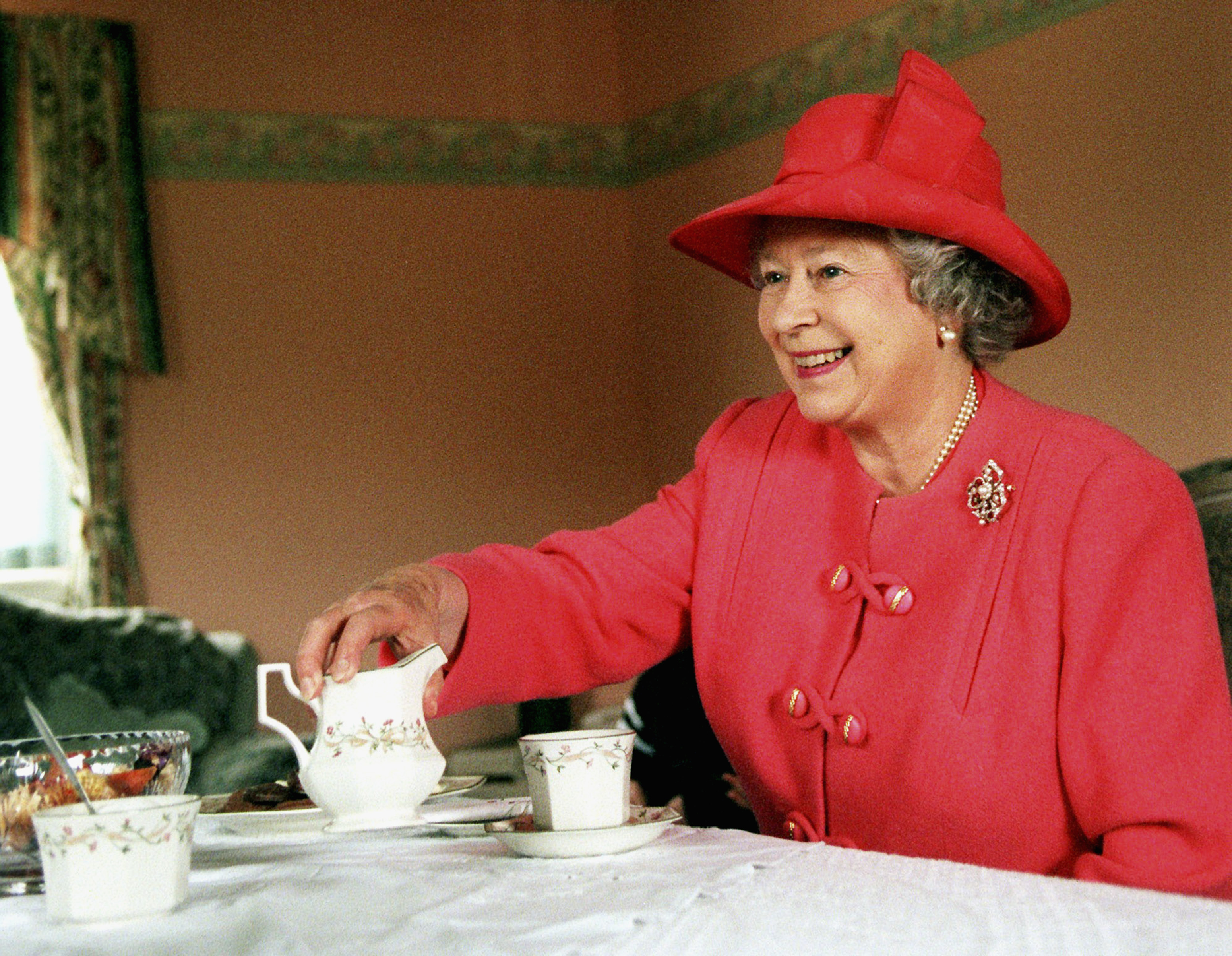 Queen Elizabeth wearing a red coat and hat drinking tea at a table