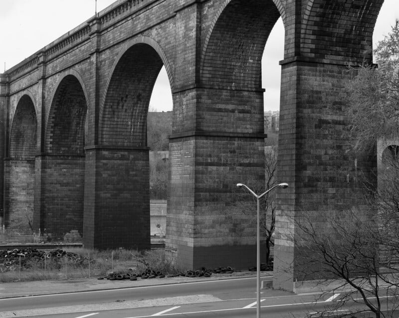 A black and white photo of a large stone railway viaduct with tall arches crossing over a road. Piles of tires are stacked near the base, and leafless trees are visible in the foreground.